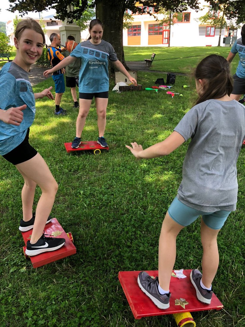 Drei Mädchen balancieren lachend auf roten Balanceboards im Gras.