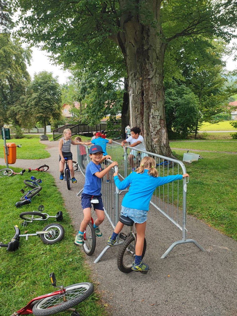 Kinder üben im Park auf Einrädern und halten sich am Haltegitter fest; ein Bub mit Kappe lacht in die Kamera.