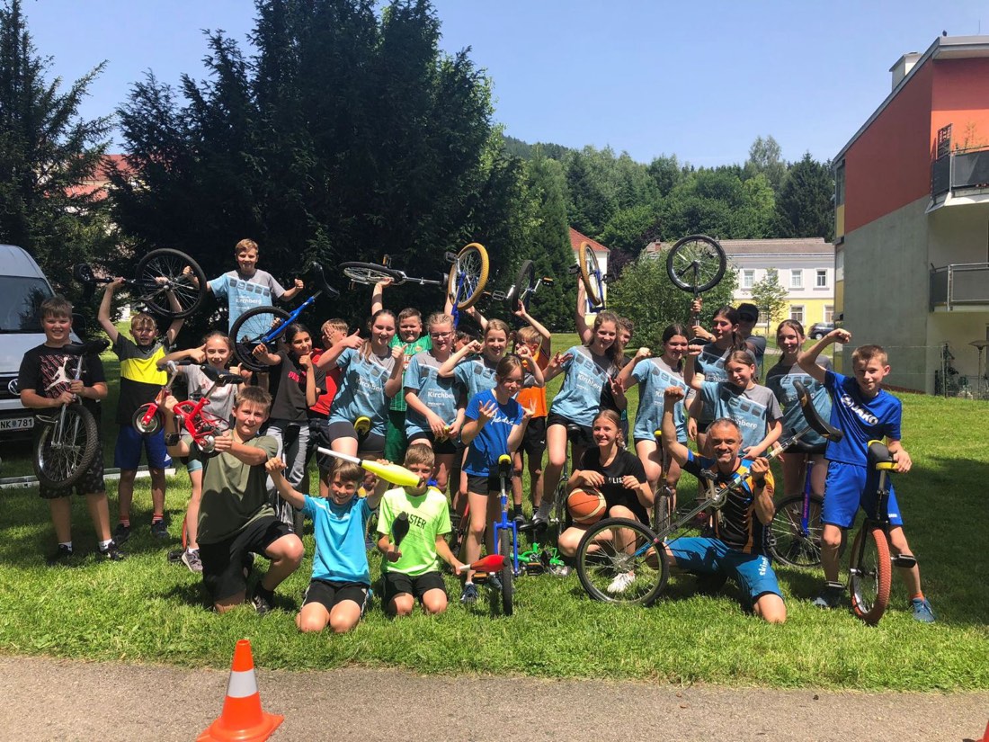 Eine ganze Schulklasse posiert jubelnd im Gras – viele Kinder halten Einräder, Jonglierkeulen und Minibikes in die Höhe, Leo kniet vorne mit einem Einrad.