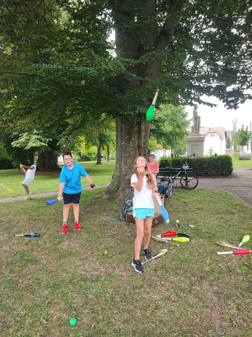 Kinder üben im Park mit bunten Jonglierkeulen; ein Mädchen schaut einer grünen Keule in der Luft nach.