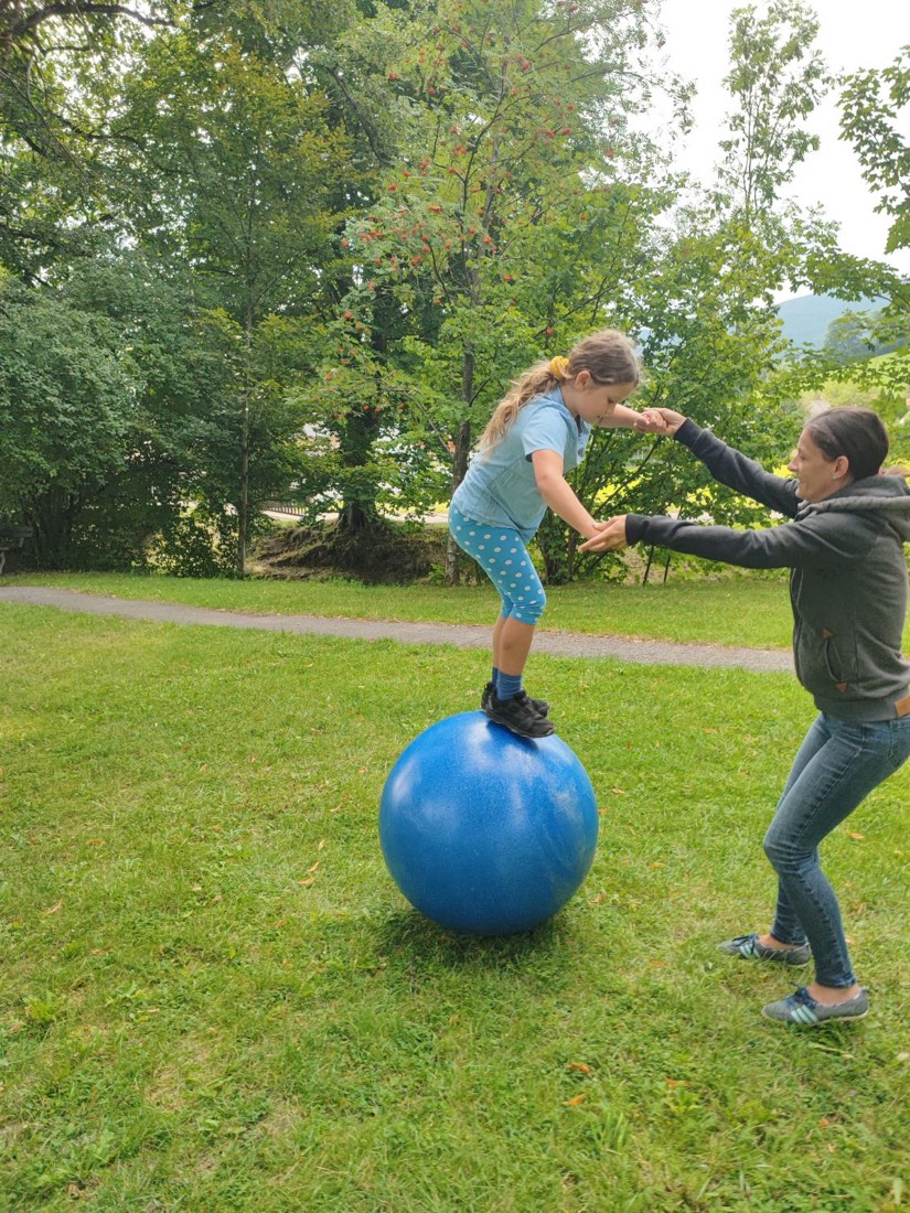 Ein Mädchen steht auf einer großen blauen Laufkugel und wird von einer Erwachsenen an den Händen gehalten.