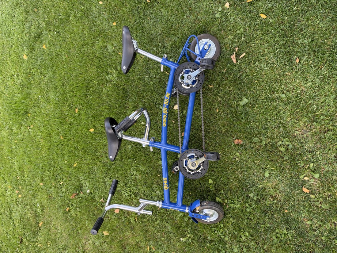 Ein blaues Mini-Tandem mit zwei Sätteln und winzigen Rädern steht im Gras.