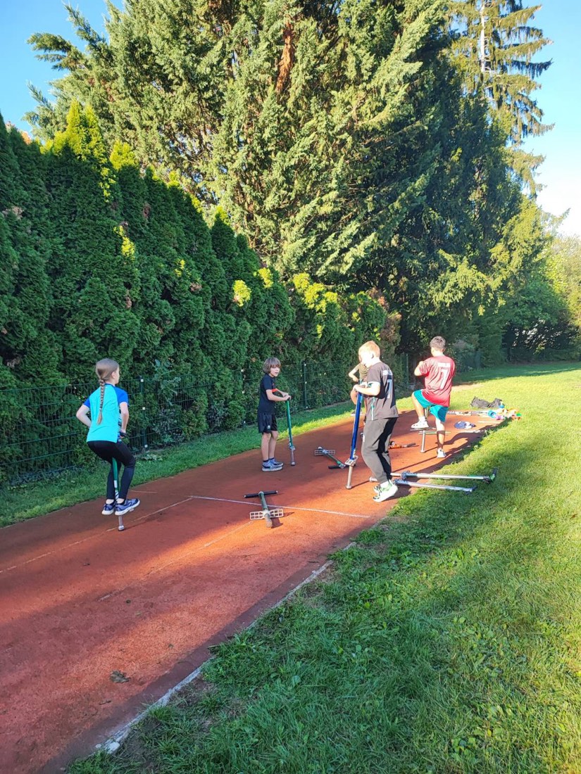 Kinder üben auf der Laufbahn mit Pogo-Sticks, weitere Pogo-Sticks liegen am Boden bereit.