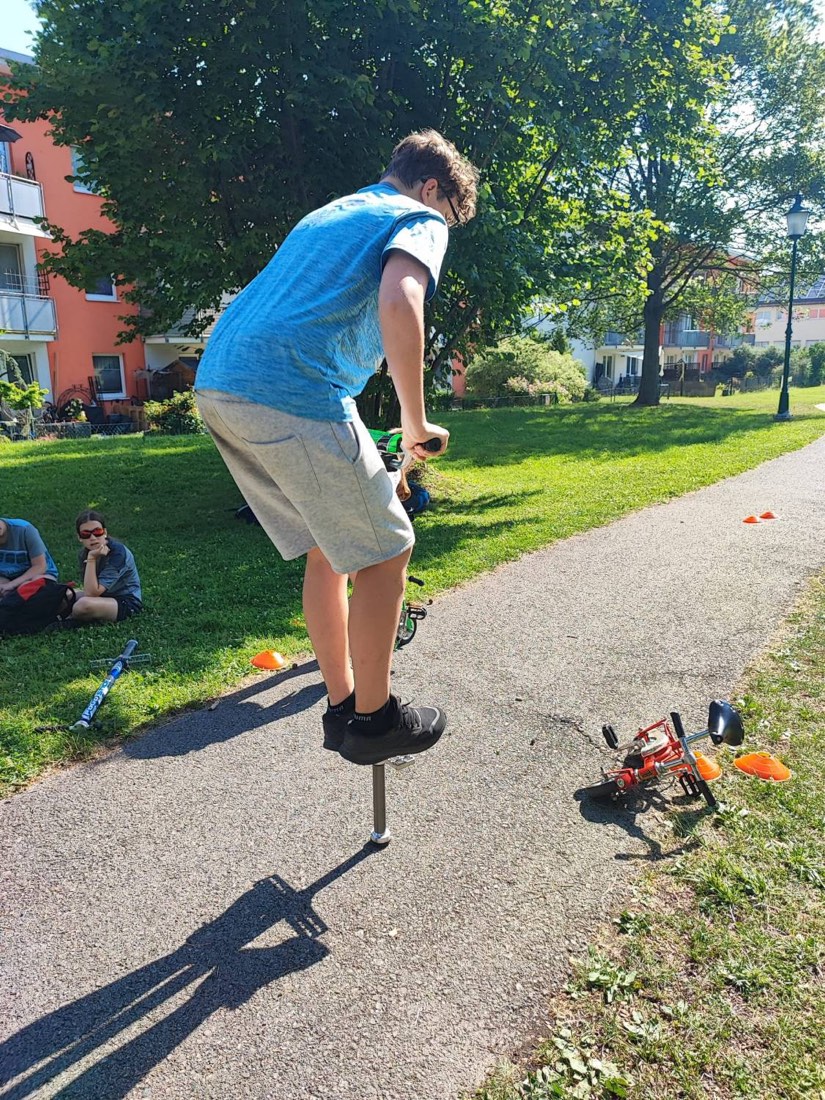 Ein Jugendlicher springt auf einem Pogo-Stick über einen Parkweg, im Gras schauen andere zu, daneben liegt ein Minibike.
