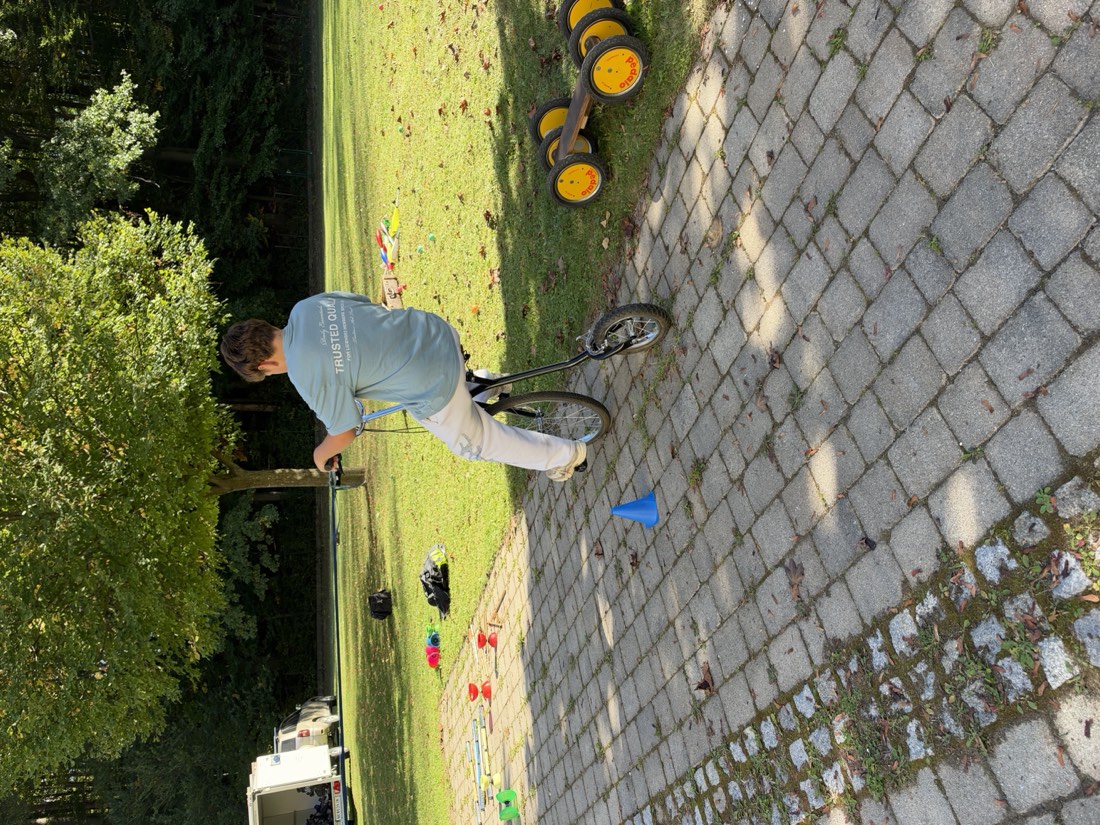 Ein Jugendlicher fährt auf einem hohen Zirkusbike über einen Pflasterweg, daneben Pedalos und Hütchen.