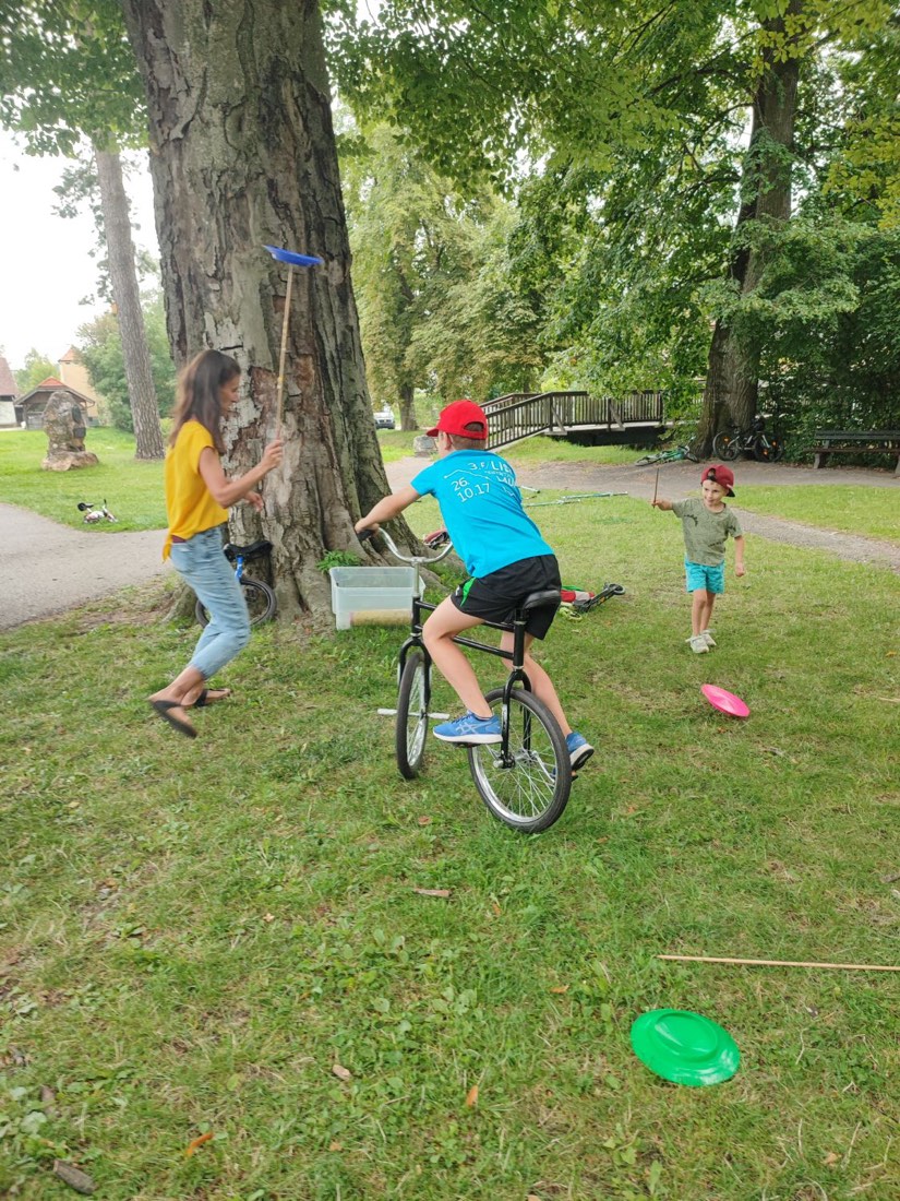 Im Park fährt ein Bub auf einem Zirkusbike, daneben drehen eine Frau und ein kleiner Bub Jonglierteller auf Stäben.