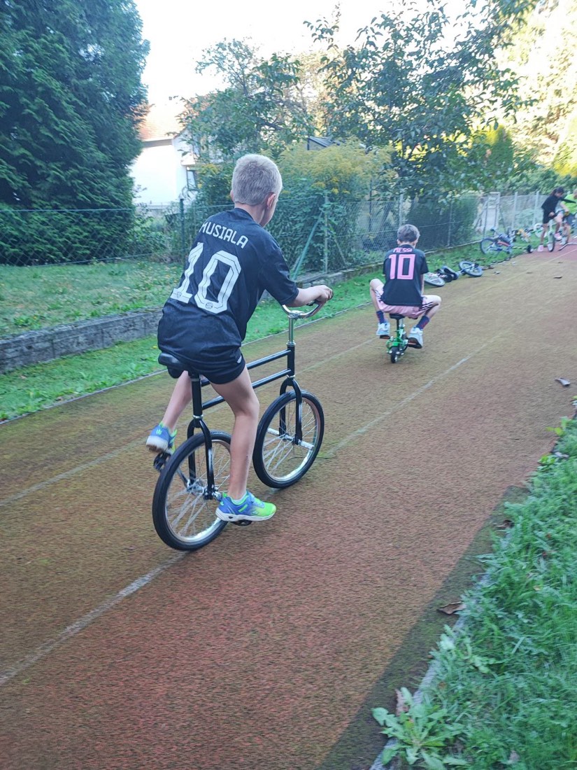 Ein Bub fährt auf einem Zirkusbike, bei dem das Vorderrad hinten ist; dahinter fährt ein Kind auf einem Minibike.
