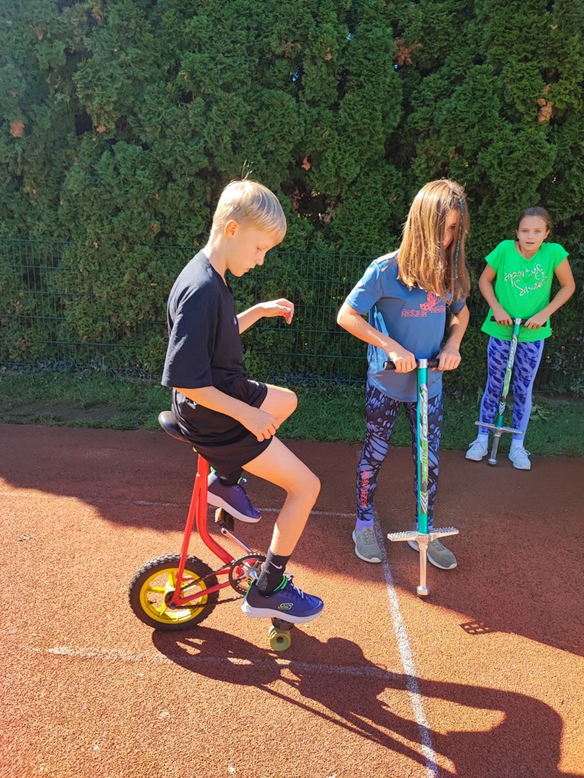 Ein Bub fährt auf einem roten Zirkusrad mit gelbem Rad, daneben üben zwei Mädchen mit Pogo-Sticks.